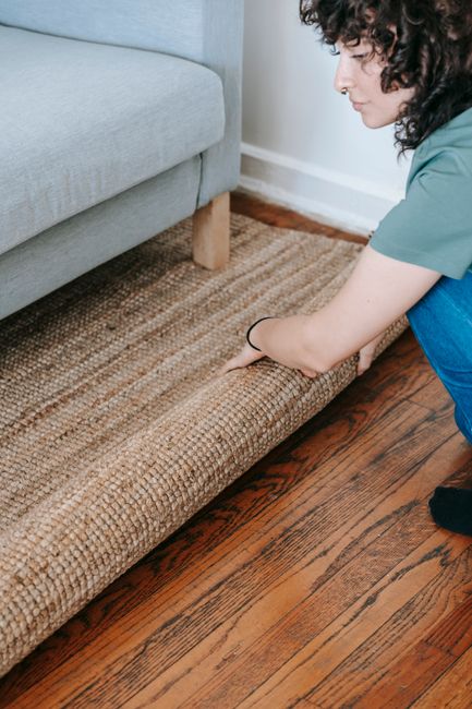 A yellow handheld vacuum cleaner connected to a flexible hose rests on an ornate patterned area rug with a floral design in a living room. The room features a hardwood floor next to the rug and has a 